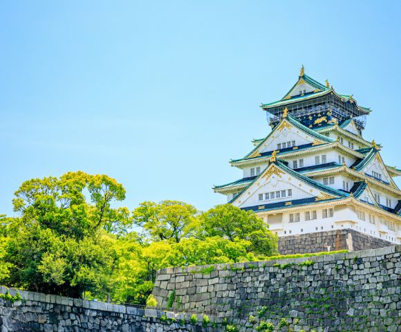 A majestic traditional Japanese castle with white walls and green tiled roofs, surrounded by lush green trees against a clear blue sky, conveys tranquility.