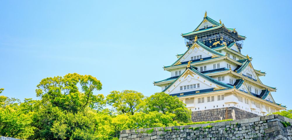 A majestic traditional Japanese castle with white walls and green tiled roofs, surrounded by lush green trees against a clear blue sky, conveys tranquility.
