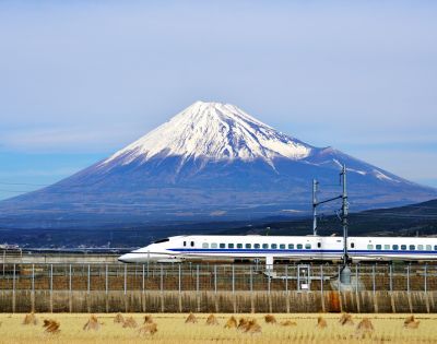 A sleek bullet train travels past golden fields with snow-capped Mount Fuji in the background under a clear blue sky, creating a serene and dynamic scene.