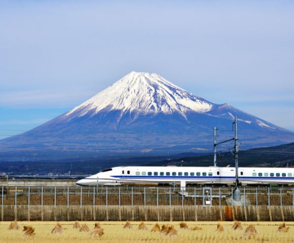 A sleek bullet train travels past golden fields with snow-capped Mount Fuji in the background under a clear blue sky, creating a serene and dynamic scene.