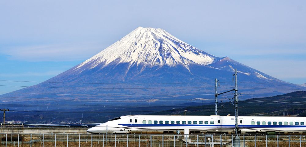 A sleek bullet train travels past golden fields with snow-capped Mount Fuji in the background under a clear blue sky, creating a serene and dynamic scene.