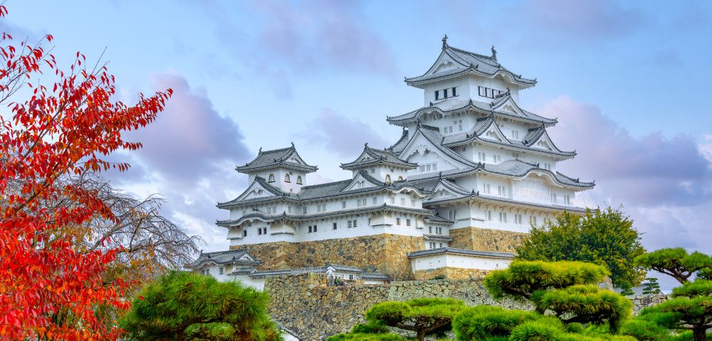 Majestic white castle with tiered roofs, surrounded by colorful autumn trees and lush green foliage, set against a blue sky with soft clouds.