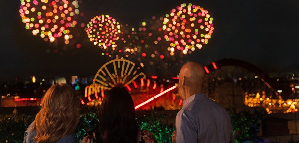 Three people watching a fireworks display at night, with colorful bursts in the sky and a view of a lit-up amusement park in the background.