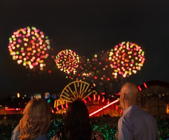 Three people watching a fireworks display at night, with colorful bursts in the sky and a view of a lit-up amusement park in the background.