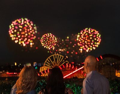 Three people watching a fireworks display at night, with colorful bursts in the sky and a view of a lit-up amusement park in the background.