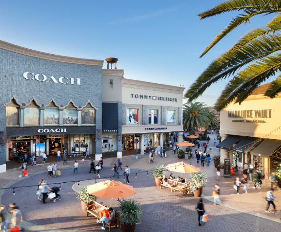 Outdoor shopping area with Coach, Tommy Hilfiger, and The Vault store facades. People walk on a sunny day, surrounded by palm trees and benches.