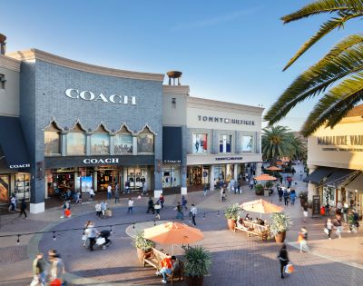 Outdoor shopping area with Coach, Tommy Hilfiger, and The Vault store facades. People walk on a sunny day, surrounded by palm trees and benches.