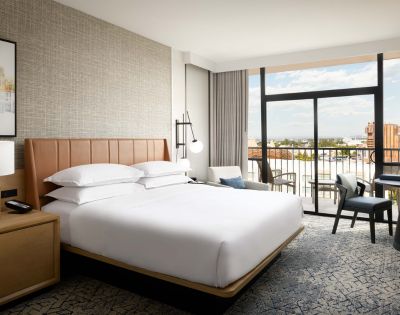 Hotel room with a large bed, modern wooden headboard, and side table with a lamp. Floor-to-ceiling windows open to a balcony with city views.