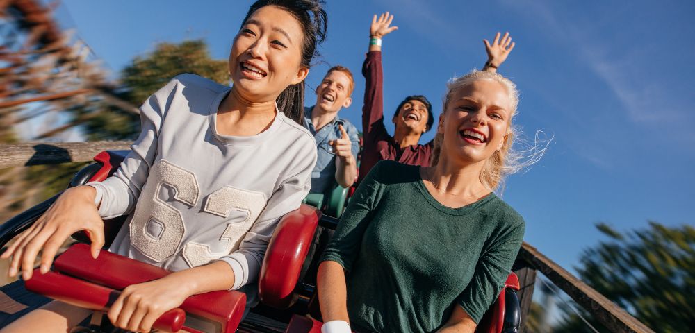 A group of friends enjoying a thrilling roller coaster ride, with hair and expressions full of excitement against a bright blue sky.