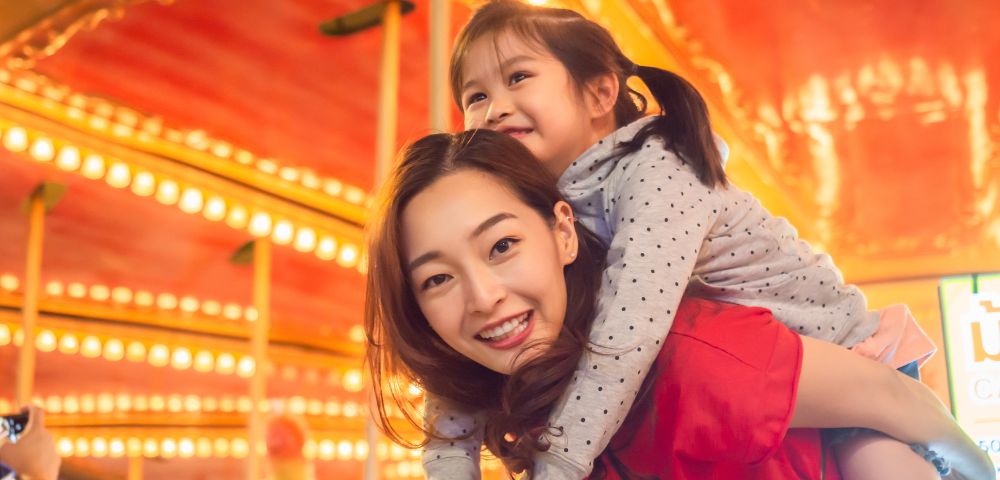 A woman carries a child on her back, smiling in front of a vibrant carnival backdrop filled with colorful lights.