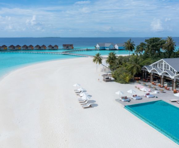 Aerial view of a serene tropical beach with white sand, turquoise water, and overwater bungalows. Palm trees surround a pool near a resort building.