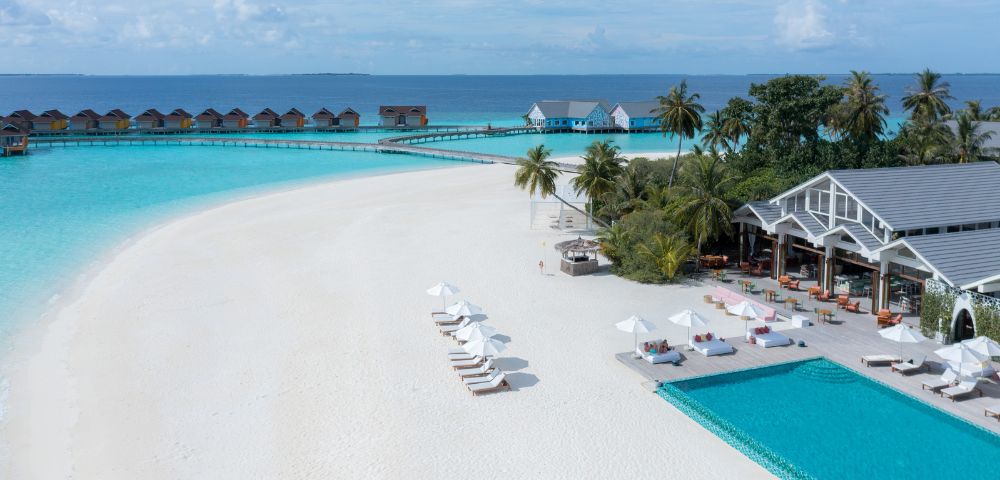 Aerial view of a serene tropical beach with white sand, turquoise water, and overwater bungalows. Palm trees surround a pool near a resort building.
