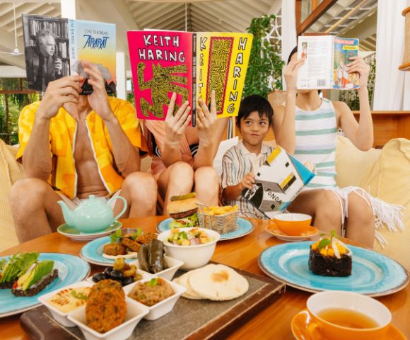 A family lounging on a sofa reads colorful books while a table in front holds an assortment of vibrant dishes and tea, creating a cozy, relaxed atmosphere.