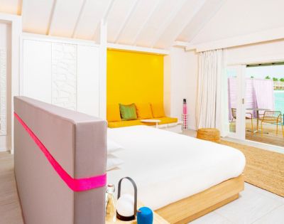 Bright, modern bedroom with a cozy bed featuring a pink accent. A vibrant yellow wall brightens the space. A window offers a view of a sunlit deck.