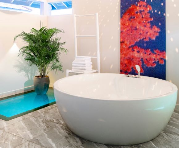 A modern bathroom with a large white oval bathtub, a tall potted plant, a white towel rack, and a vibrant red-blue abstract painting on the wall.