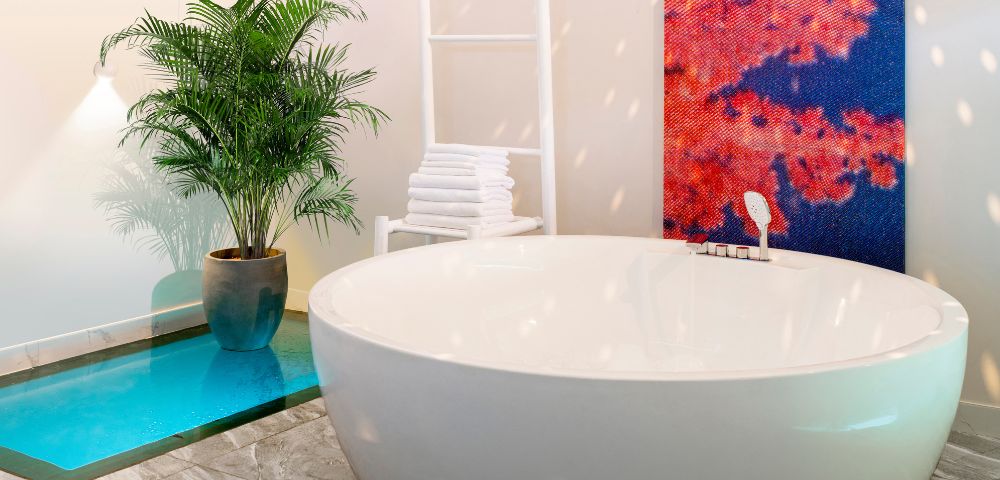 A modern bathroom with a large white oval bathtub, a tall potted plant, a white towel rack, and a vibrant red-blue abstract painting on the wall.