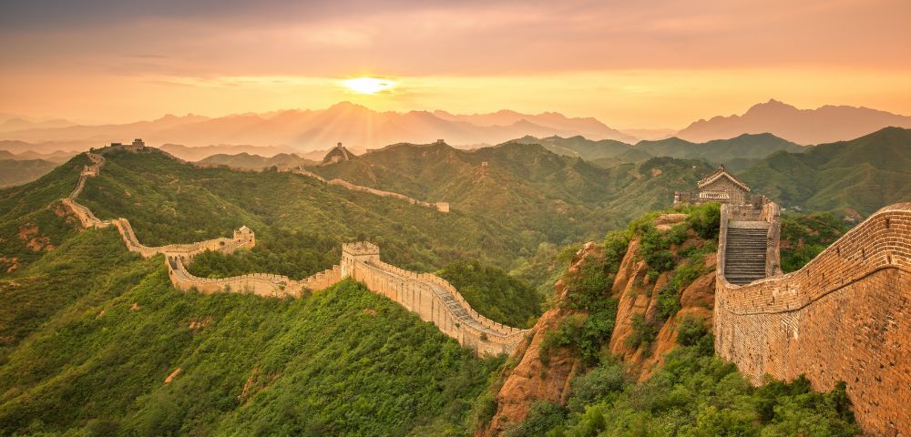 Sunset view of the Great Wall of China meandering over lush green hills, with the sun casting a warm glow over distant mountains, evoking tranquility and grandeur.