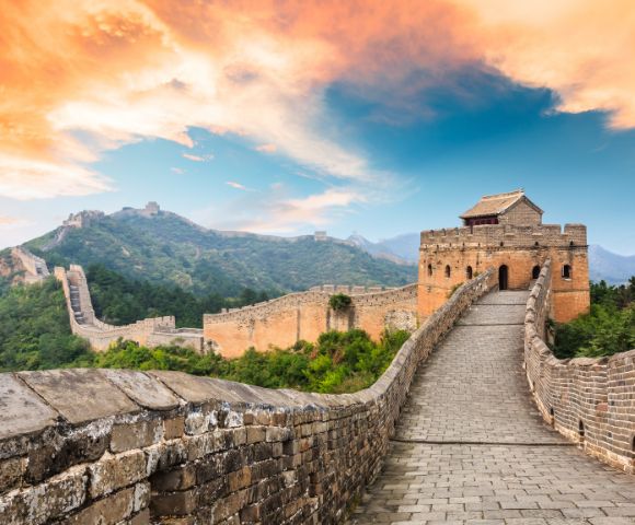 The Great Wall of China stretches over lush green hills under a vibrant orange and blue sky. A prominent tower stands in the foreground, evoking awe.