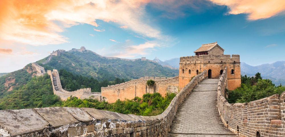 The Great Wall of China stretches over lush green hills under a vibrant orange and blue sky. A prominent tower stands in the foreground, evoking awe.