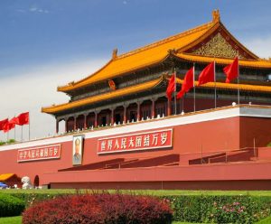 Historical building with ornate red walls and an intricate, golden roof under a clear blue sky. Red flags line the structure, with lush greenery nearby.