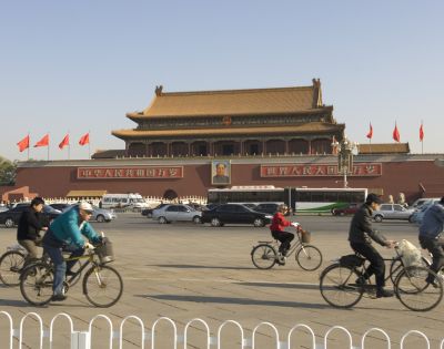 Cyclists ride past the Tiananmen Gate in Beijing under a clear sky. The historic building is adorned with red flags and a prominent portrait.