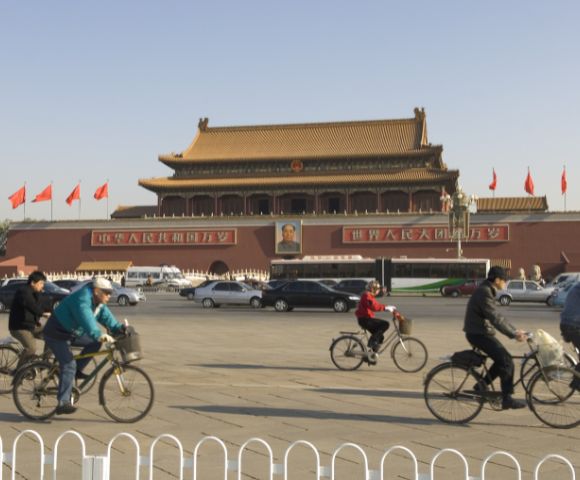 Cyclists ride past the Tiananmen Gate in Beijing under a clear sky. The historic building is adorned with red flags and a prominent portrait.