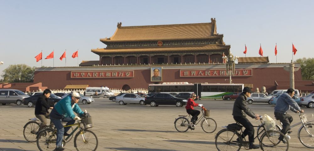 Cyclists ride past the Tiananmen Gate in Beijing under a clear sky. The historic building is adorned with red flags and a prominent portrait.