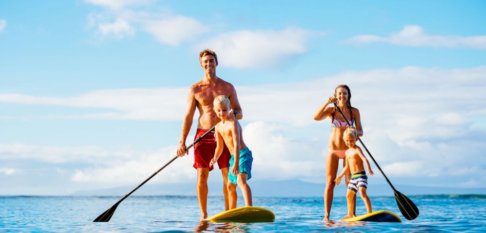 A family of four, including a father, mother, and two young children, paddleboarding together on a calm, sunlit ocean under a blue sky. They each stand on their own paddleboard, and are holding paddles.