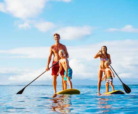 A family of four, including a father, mother, and two young children, paddleboarding together on a calm, sunlit ocean under a blue sky. They each stand on their own paddleboard, and are holding paddles.