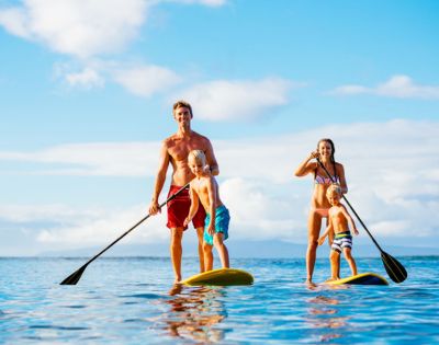 A family of four, including a father, mother, and two young children, paddleboarding together on a calm, sunlit ocean under a blue sky. They each stand on their own paddleboard, and are holding paddles.