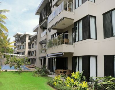 Modern apartment complex with balconies and glass railings, surrounded by lush greenery and palm trees. A pool is visible in the background.