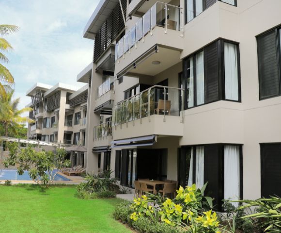 Modern apartment complex with balconies and glass railings, surrounded by lush greenery and palm trees. A pool is visible in the background.