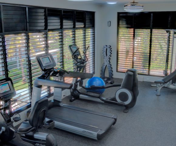 Modern gym room with cardio machines, a treadmill, and a recumbent bike by large windows with blinds. Bright, spacious, and inviting atmosphere.