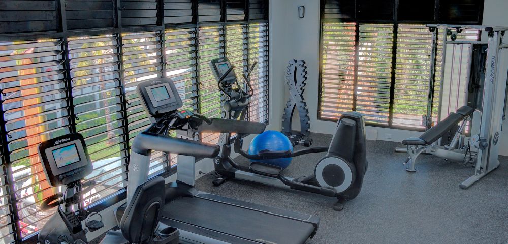 Modern gym room with cardio machines, a treadmill, and a recumbent bike by large windows with blinds. Bright, spacious, and inviting atmosphere.