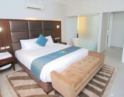 Modern hotel room with a king-size bed, brown cushioned headboard, and blue accents. A tan bench sits at the foot. Bright and inviting atmosphere.