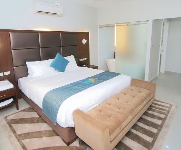 Modern hotel room with a king-size bed, brown cushioned headboard, and blue accents. A tan bench sits at the foot. Bright and inviting atmosphere.