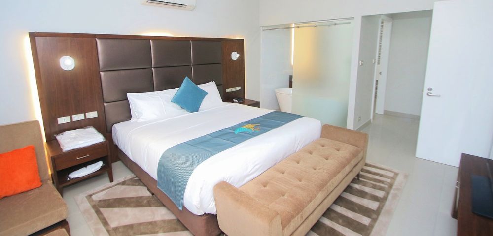 Modern hotel room with a king-size bed, brown cushioned headboard, and blue accents. A tan bench sits at the foot. Bright and inviting atmosphere.