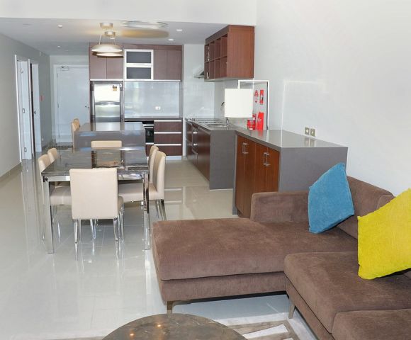 Modern living room with a brown sectional sofa adorned with blue and yellow pillows. The open kitchen and dining area feature sleek cabinetry, a stainless steel fridge, and beige chairs around a glossy table, conveying a cozy, contemporary ambiance.