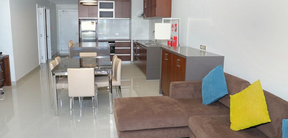 Modern living room with a brown sectional sofa adorned with blue and yellow pillows. The open kitchen and dining area feature sleek cabinetry, a stainless steel fridge, and beige chairs around a glossy table, conveying a cozy, contemporary ambiance.