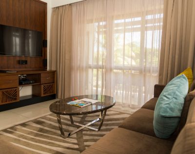 A cozy living room with a brown sofa, blue and yellow cushions, a marble coffee table, and an entertainment center with a flat-screen TV. Beige curtains filter soft sunlight.