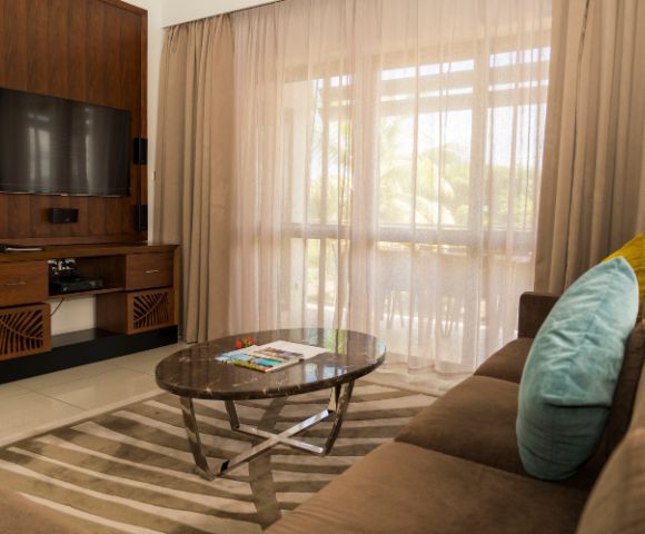 A cozy living room with a brown sofa, blue and yellow cushions, a marble coffee table, and an entertainment center with a flat-screen TV. Beige curtains filter soft sunlight.