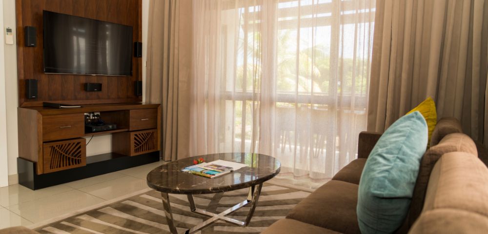A cozy living room with a brown sofa, blue and yellow cushions, a marble coffee table, and an entertainment center with a flat-screen TV. Beige curtains filter soft sunlight.