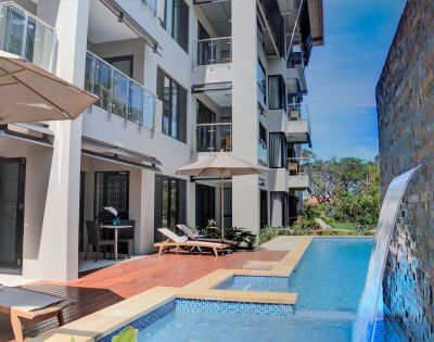 Modern apartment complex with a poolside view. A cascading waterfall, sun loungers, and umbrellas create a relaxed tropical ambiance.