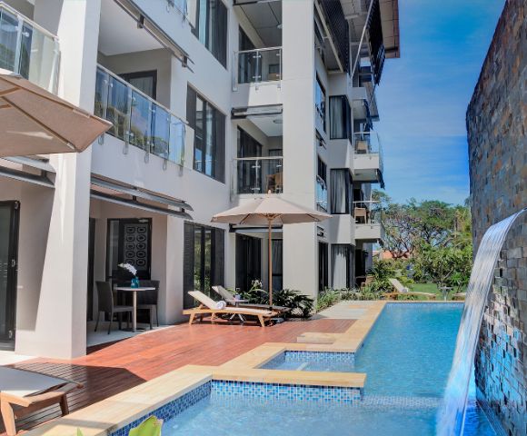 Modern apartment complex with a poolside view. A cascading waterfall, sun loungers, and umbrellas create a relaxed tropical ambiance.