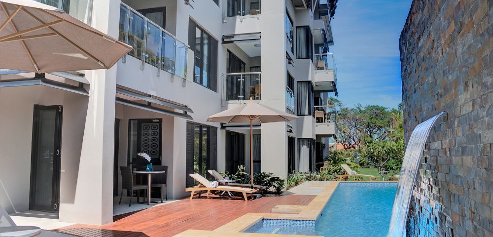 Modern apartment complex with a poolside view. A cascading waterfall, sun loungers, and umbrellas create a relaxed tropical ambiance.