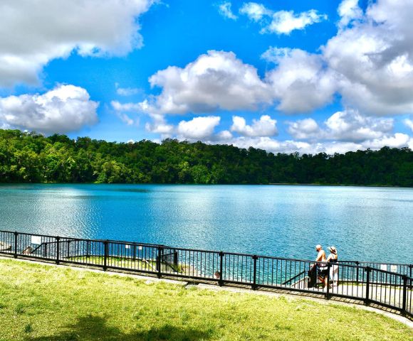 A tranquil lake surrounded by lush green forest under a bright blue sky with fluffy clouds. A couple stands by a railing, enjoying the serene view.