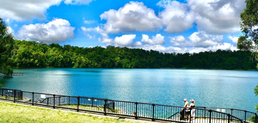 A tranquil lake surrounded by lush green forest under a bright blue sky with fluffy clouds. A couple stands by a railing, enjoying the serene view.
