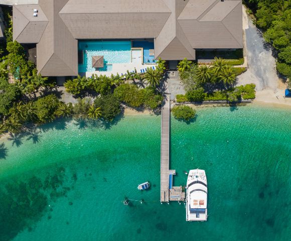 Aerial view of a tropical resort with a large building, pool, and lush greenery. A wooden dock extends to a white yacht on clear turquoise water.
