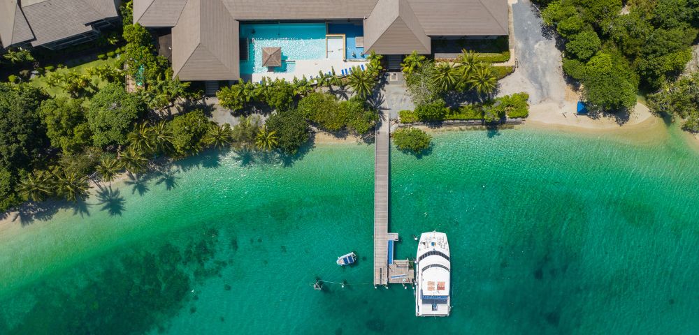 Aerial view of a tropical resort with a large building, pool, and lush greenery. A wooden dock extends to a white yacht on clear turquoise water.