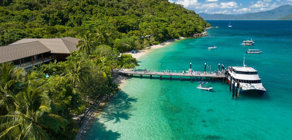 Tropical beach scene with lush green forest, turquoise water, and a dock with a catamaran and boats. Visitors stroll under a sunny blue sky.